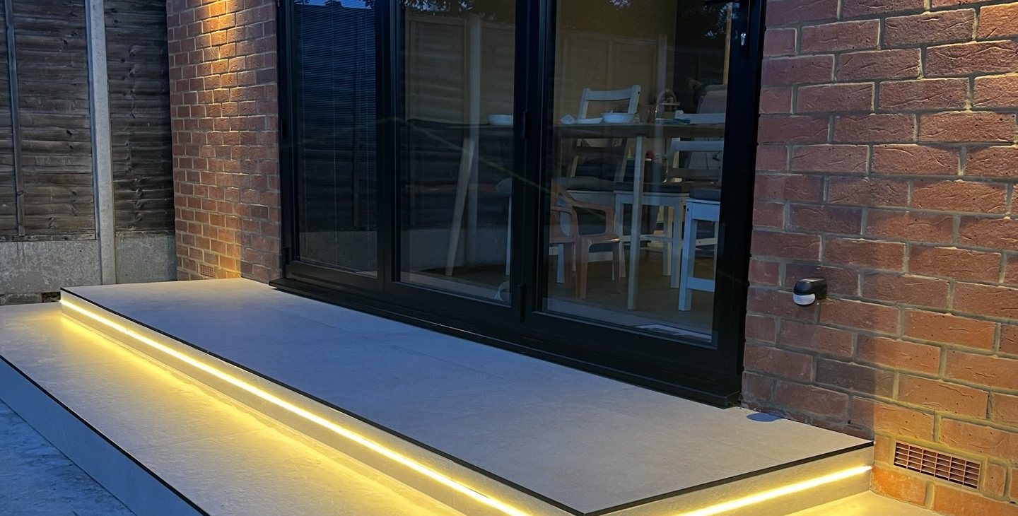 The finished rear extension at dusk, black bifold doors and a lit step onto the terrace, Bexleyheath DA7